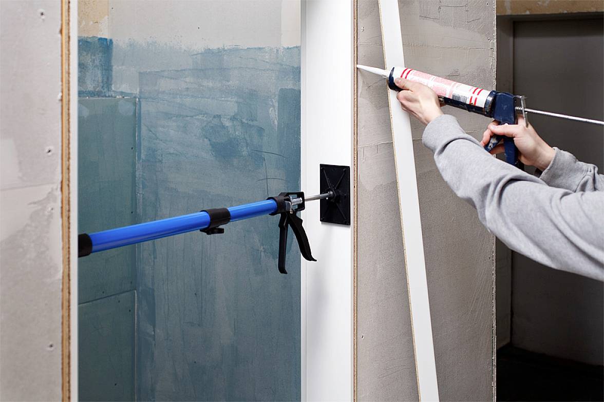 Eine Person befestigt mit einer Dichtpistole eine weiße Leiste an der Kante einer Wand. Eine Klemme hält die Leiste in Position.