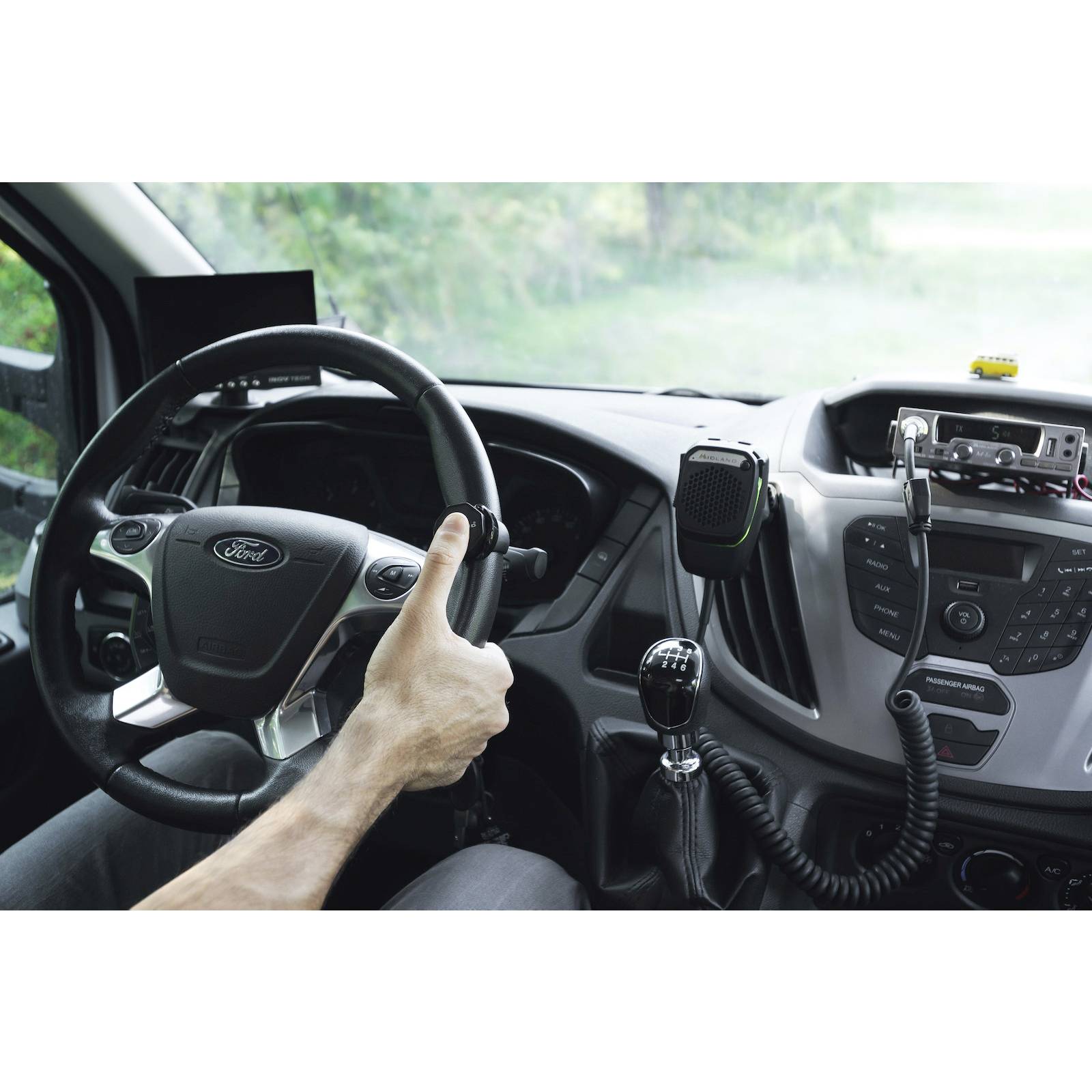 Ein Lenkrad in einem Auto, mit elektronischen Geräten an der Armaturentafel. Eine Hand hält das Lenkrad. Im Hintergrund ist Grün zu sehen.