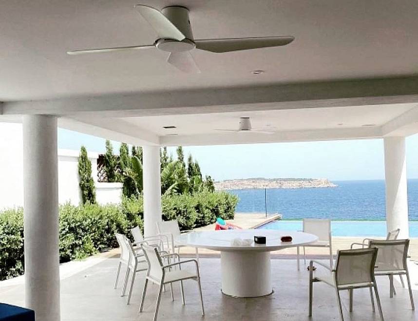 Überdachte Terrasse mit weißen Stühlen und rundem Tisch, Blick auf blaues Meer und Küste, Pool im Hintergrund, sonniger Tag.