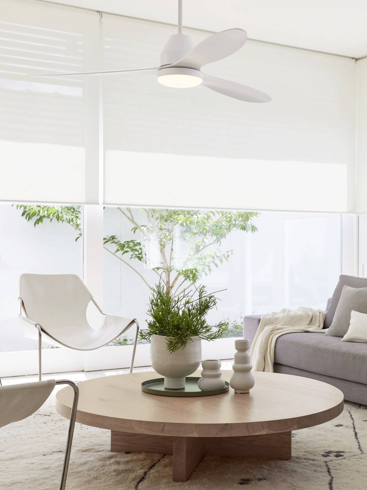 Helles, modernes Wohnzimmer mit rundem Holztisch, weißem Stuhl, grauem Sofa und Zimmerpflanze; große Fenster im Hintergrund.