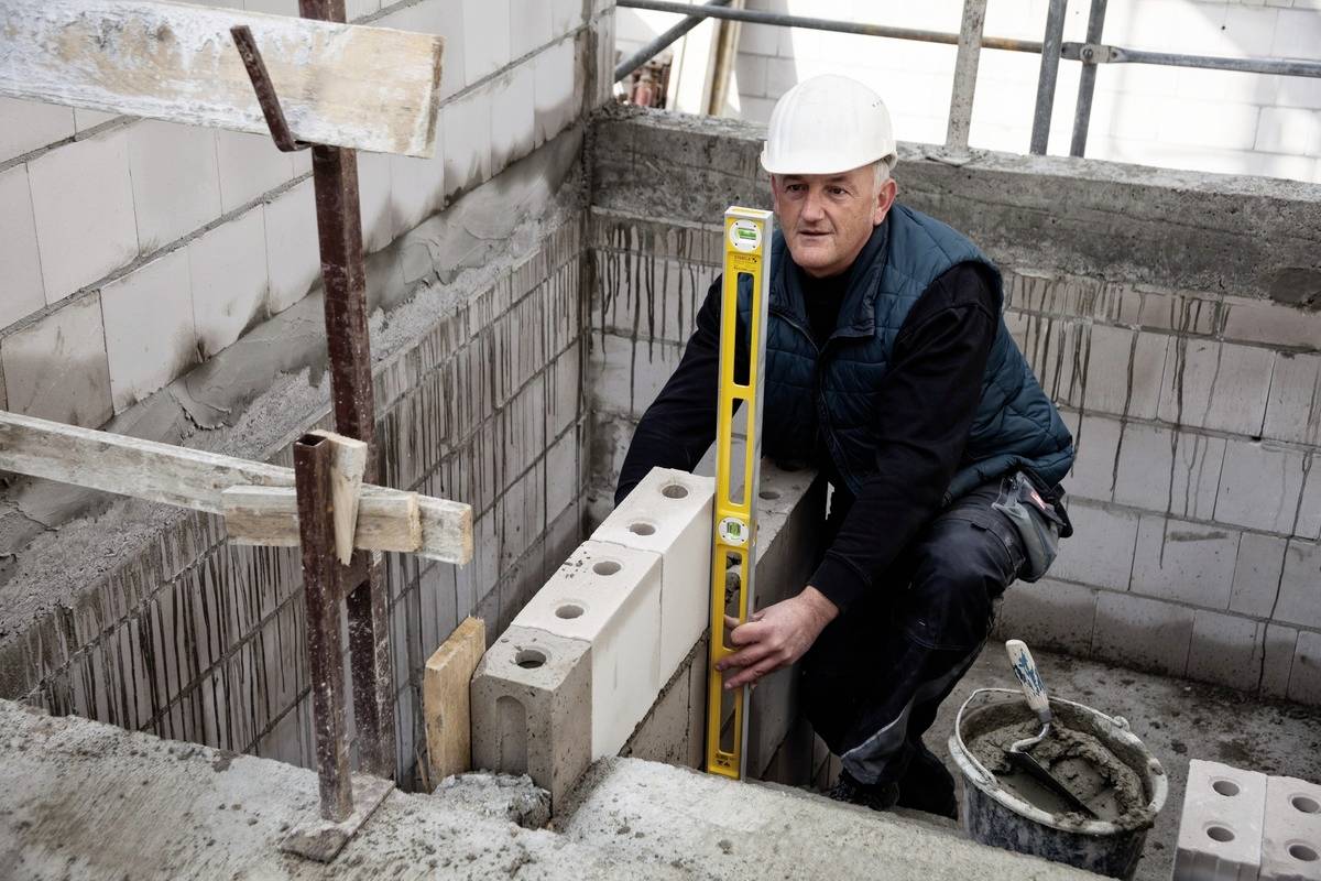 Ein Bauarbeiter mit Schutzhelm verwendet eine Wasserwaage, um eine Ziegelwand gerade zu errichten. Baustelle mit Gerüsten im Hintergrund.
