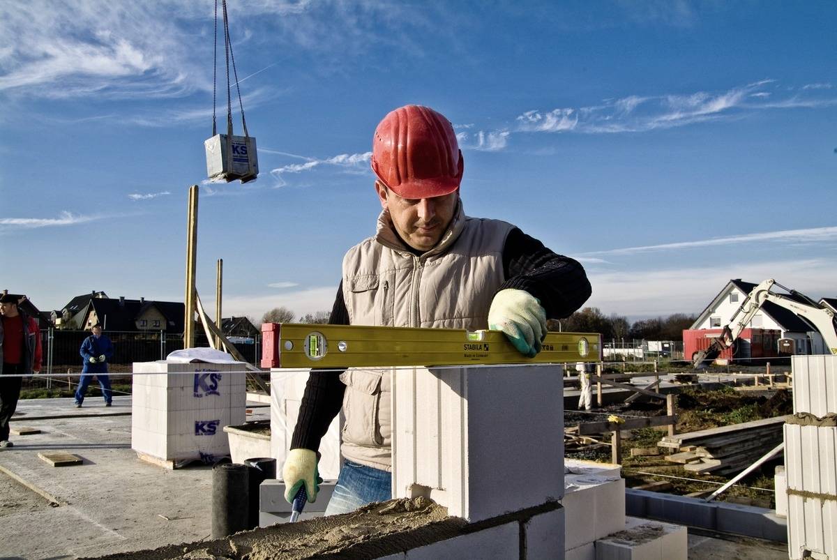 Ein Bauarbeiter mit rotem Helm und Handschuhen richtet Ziegel an einer Baustelle aus, verwendet eine Wasserwaage, blauer Himmel im Hintergrund.