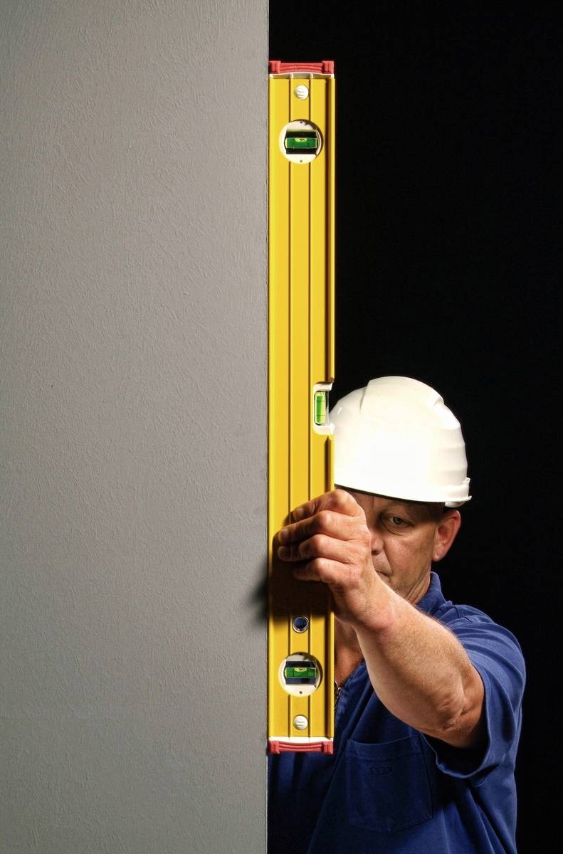 Ein Bauarbeiter mit einem weißen Helm überprüft mit einer Wasserwaage die Vertikalität einer grauen Wand.