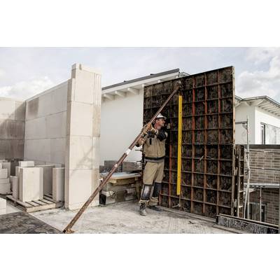 Ein Bauarbeiter errichtet eine Wand auf einer Baustelle. Er trägt Schutzkleidung und verwendet eine Stütze, um die Wand abzusichern. Im Hintergrund sind weitere Gebäude zu sehen.