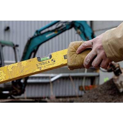 'Nahaufnahme einer Hand, die eine Wasserwaage hält; im Hintergrund ein Bagger auf einer Baustelle.'