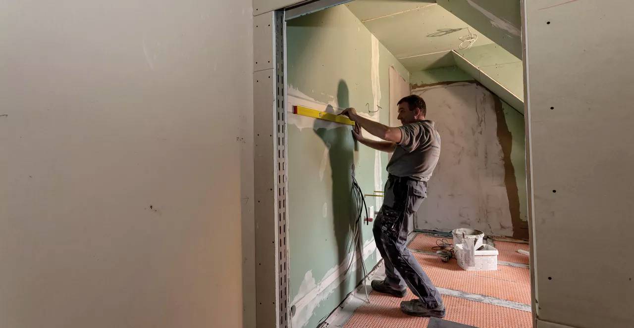 Ein Handwerker prüft mit einer Wasserwaage die Ausrichtung einer frisch installierten Trockenbauwand in einem unfertigen Raum.