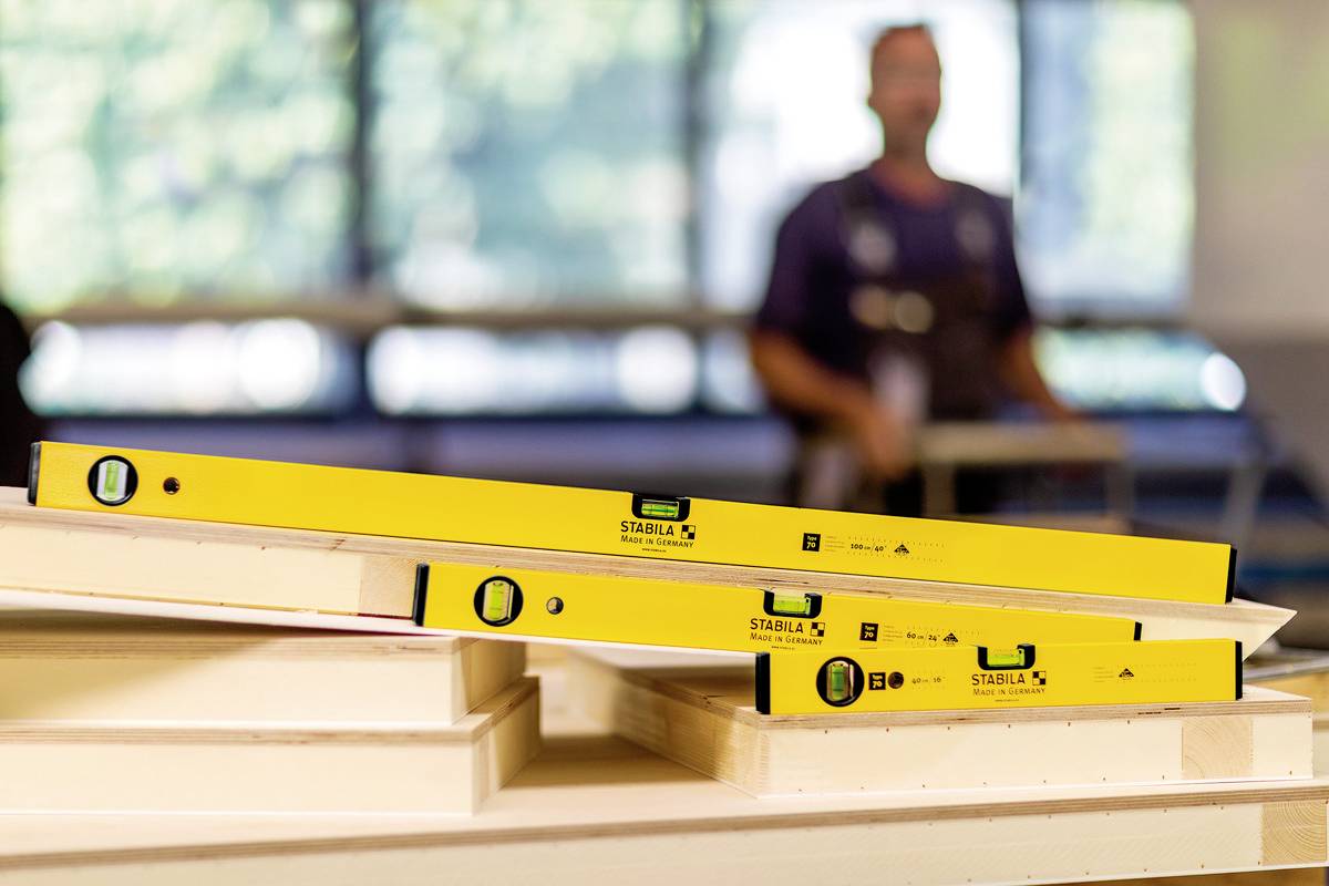 Gelbe Wasserwaagen liegen auf Holzplatten, im Hintergrund steht unscharf eine Person in einem Workshop oder Baubereich.