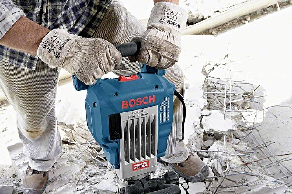 Eine Person arbeitet mit einem blauen Bosch Abbruchhammer auf Beton, trägt Handschuhe, zerbröckelte Betonteile und freiliegende Metallstäbe sichtbar.
