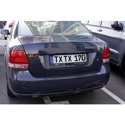Ein dunkles Auto steht auf einem Parkplatz; das Kennzeichen lautet 'TXTX 170'. Im Hintergrund sind hohe Gebäude zu sehen.