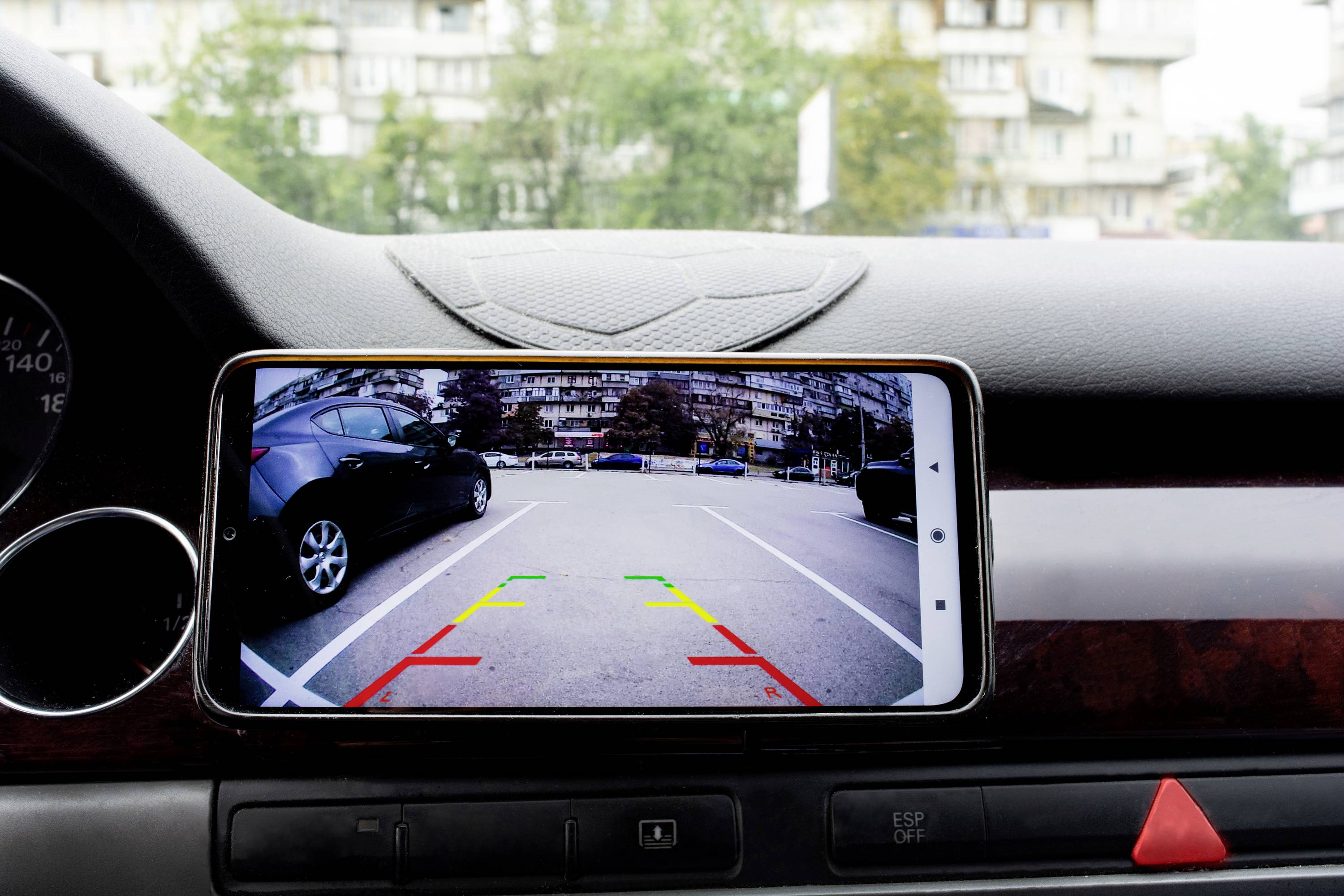 Ein Smartphone zeigt auf einem Armaturenbrett den Rückfahrkamera-Bildschirm mit farbigen Linien zur Einparkhilfe in einem Parkplatz.