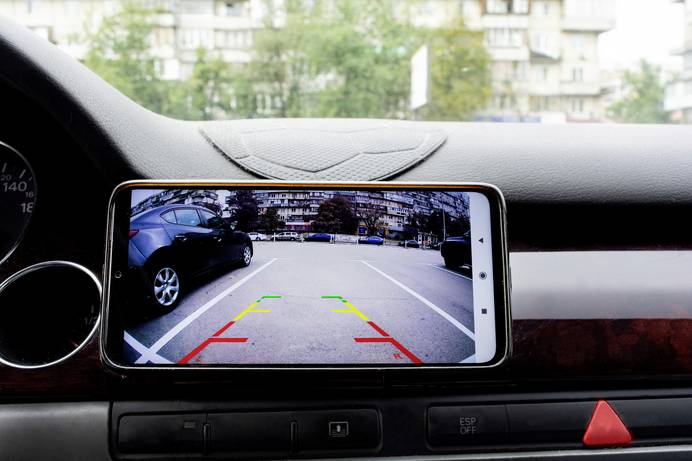 Man sieht ein Smartphone, das im Cockpit eines Autos angebracht ist und als Bildschirm für die Bildübertragung der Rückfahrkamera dient. Auf dem Monitor sind ein leerer Parkplatz und farbige Hilfslinien, die beim Rückwärtseinparken helfen sollen, zu sehen.