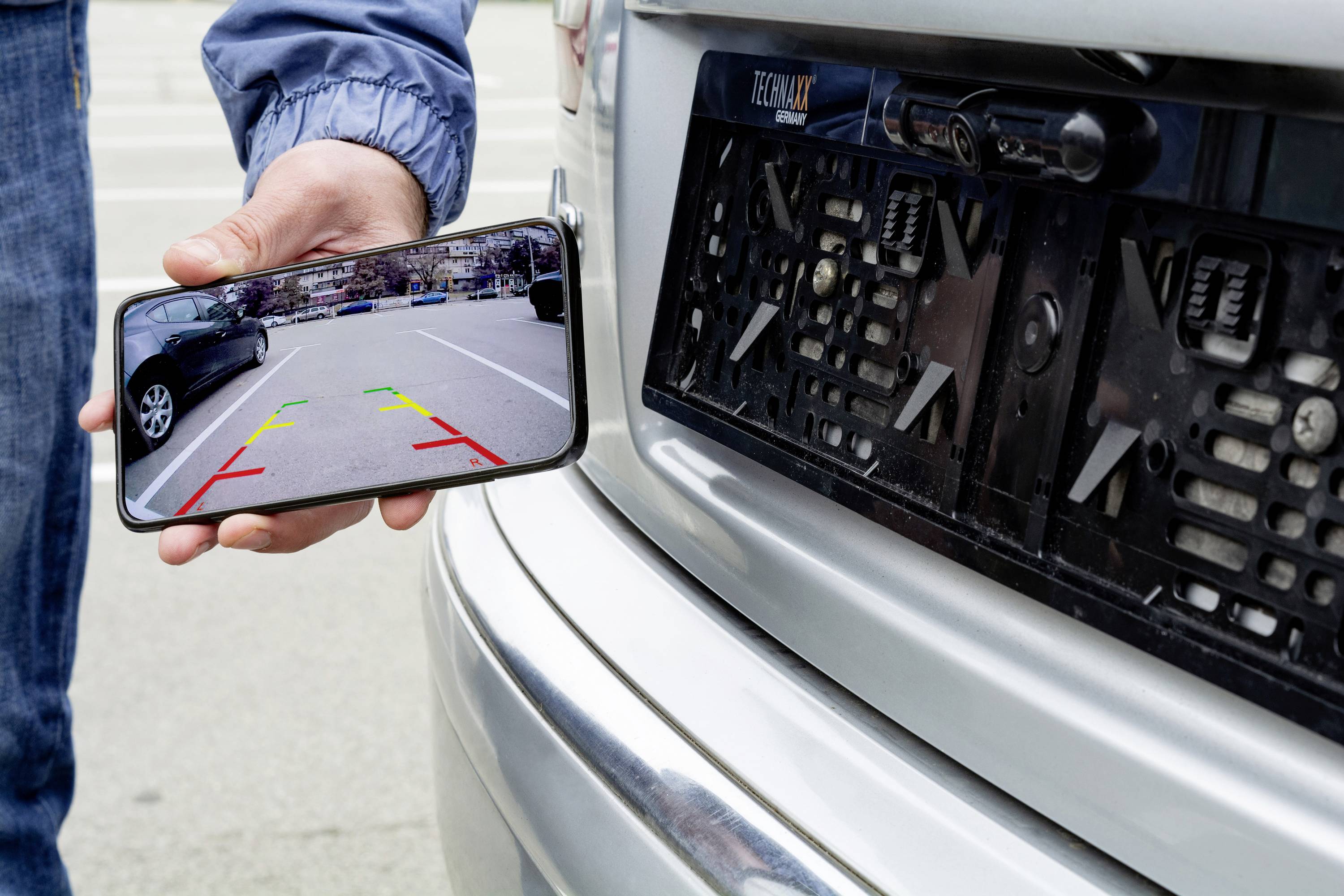 Ein Smartphone zeigt das Bild einer Rückfahrkamera mit farbigen Linien, während es an das Heck eines Autos gehalten wird.