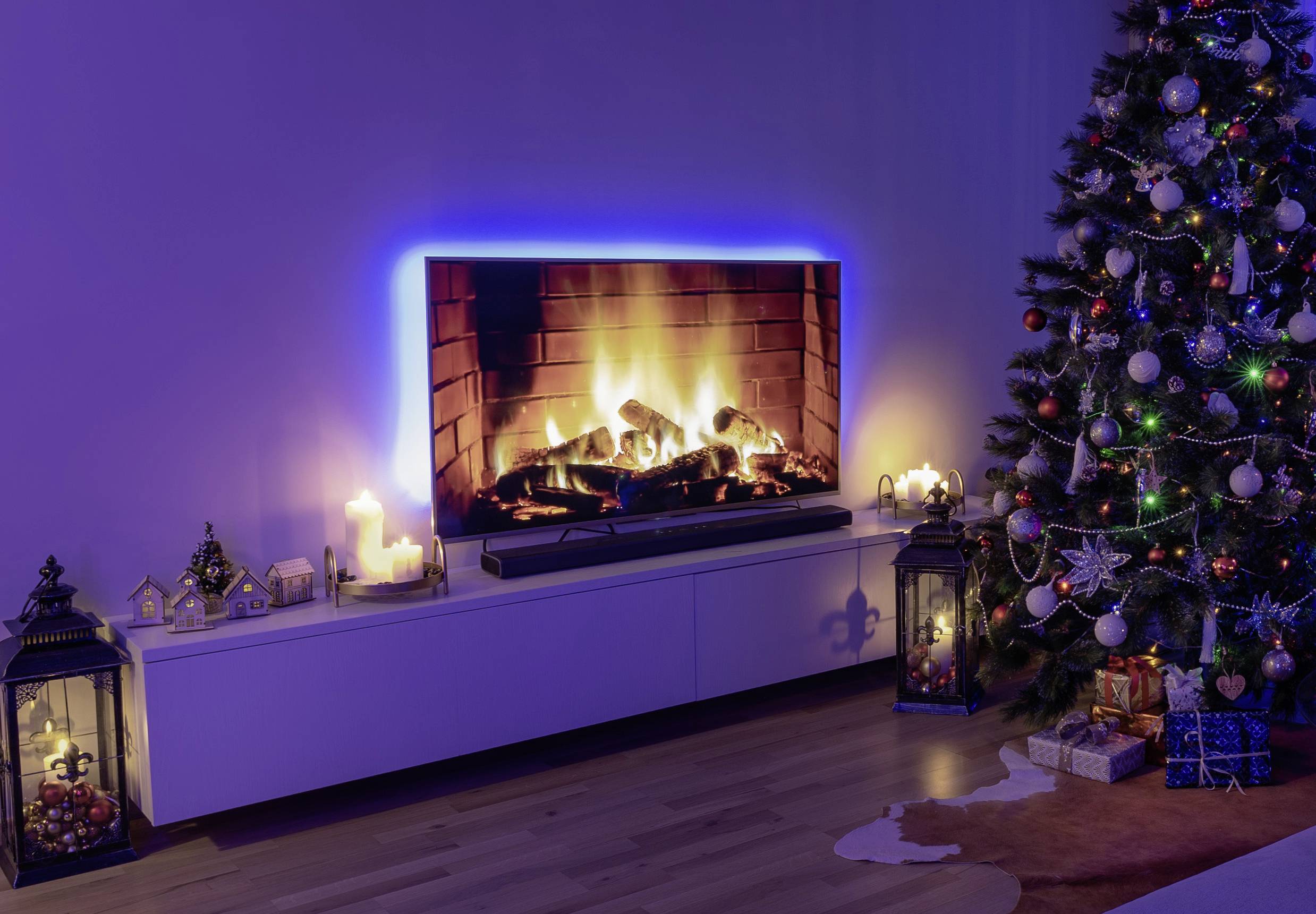 Ein Wohnzimmer mit einem Fernseher, der ein Kaminfeuer zeigt, umgeben von Kerzen und Weihnachtsschmuck. Rechts steht ein geschmückter Weihnachtsbaum.