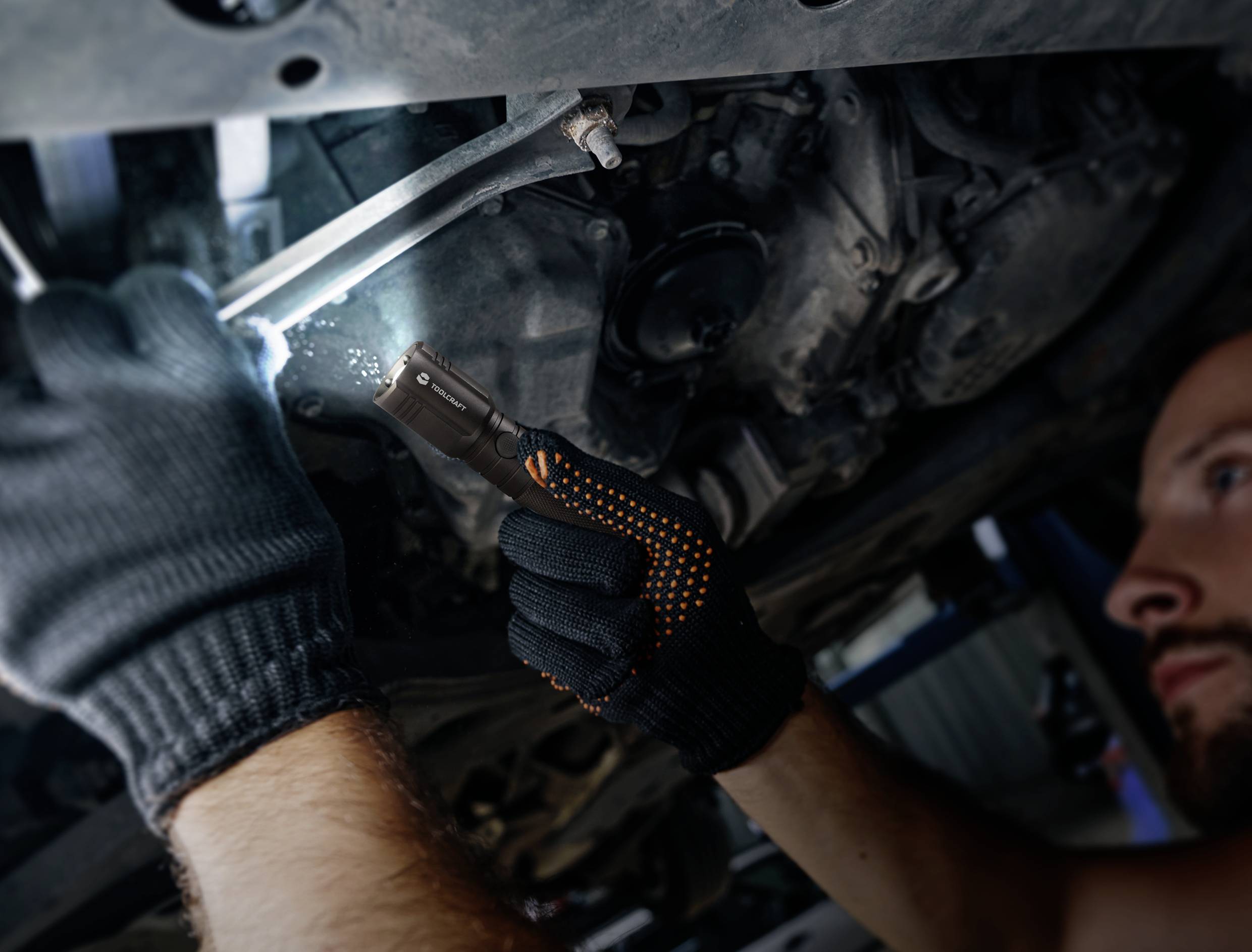 Eine Person repariert mit Handschuhen und Lampe ein Auto unter der Motorhaube, fokusiert und arbeitet an Motorteilen.