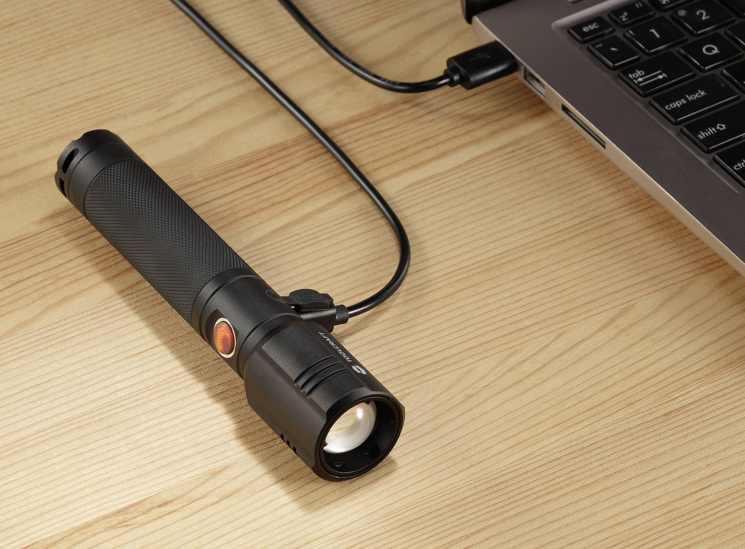 Eine Taschenlampe ist mit einem Ladekabel an einen Laptop auf einem Holztisch angeschlossen.