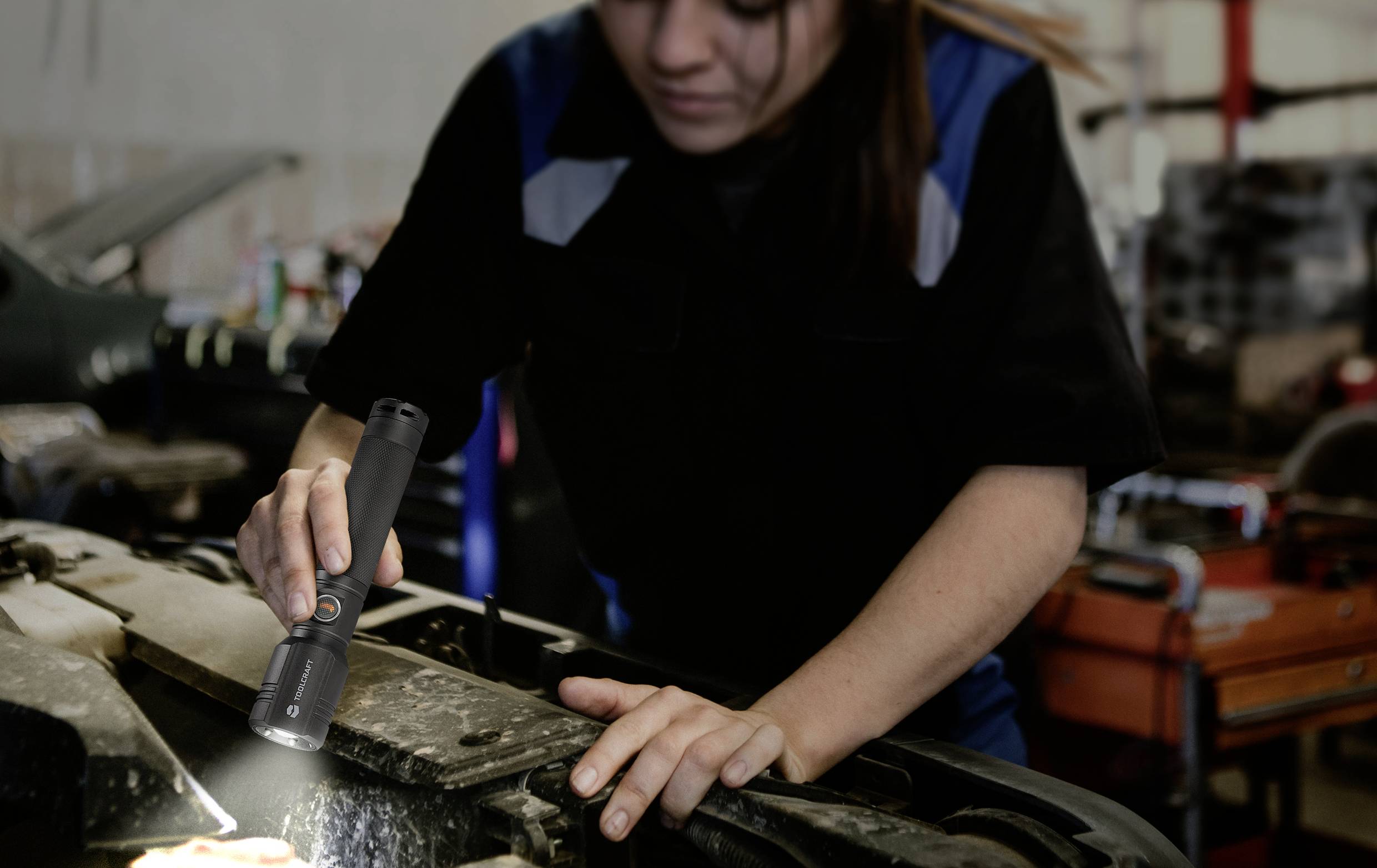 Eine Mechanikerin inspiziert mit einer Taschenlampe den Motorraum eines Autos in einer Werkstatt.