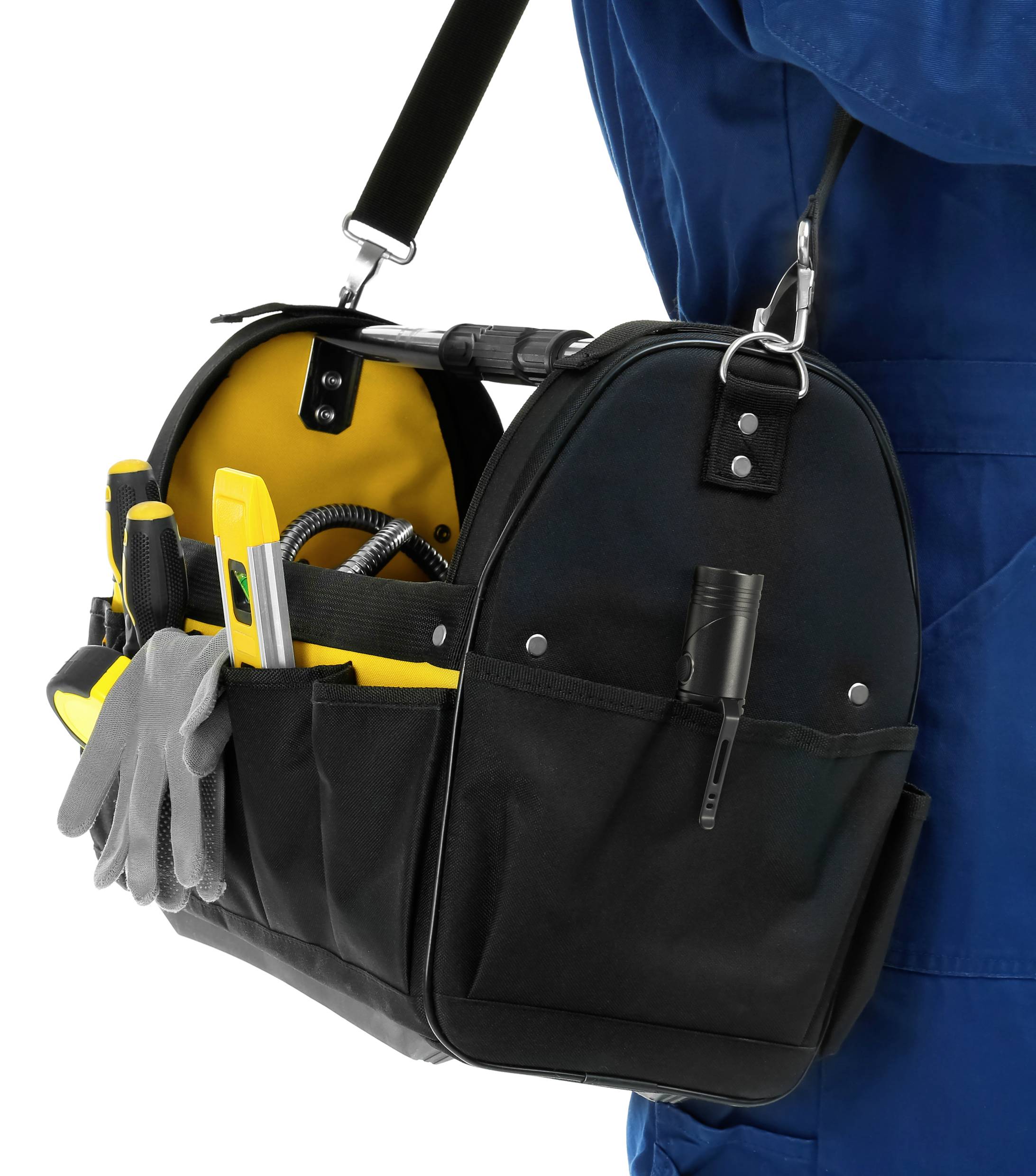 Eine schwarze Werkzeugtasche mit gelben Details, gefüllt mit verschiedenen Werkzeugen, hängt an der Schulter eines Mannes in blauer Arbeitskleidung.