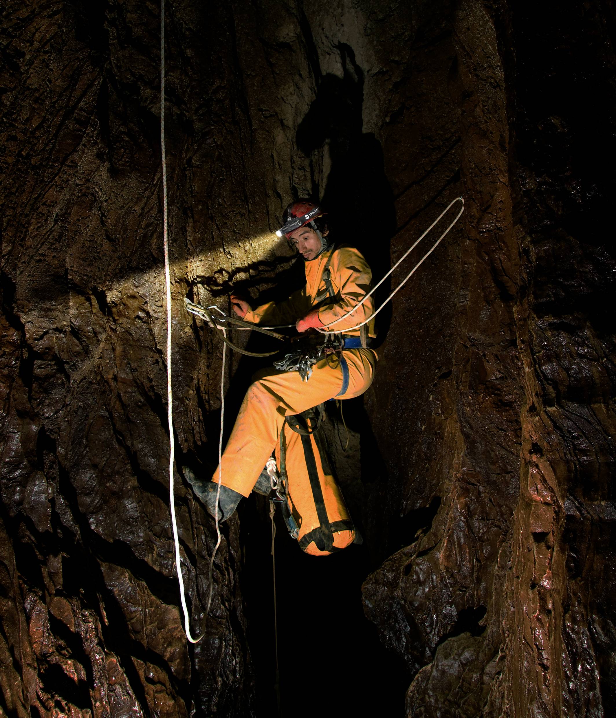 Eine Person in Kletterausrüstung steigt in einer dunklen Höhle mit Seilen hinab. Helm mit Stirnlampe beleuchtet den felsigen Hintergrund.