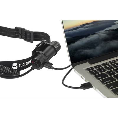 Eine Stirnlampe der Marke Toolcraft ist per Kabel mit einem Laptop verbunden, auf dem ein Sonnenaufgang über Wolken zu sehen ist.