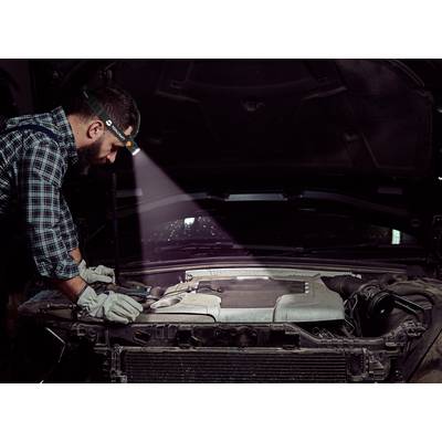Ein Mechaniker mit Stirnlampe arbeitet unter einer geöffneten Motorhaube. Er trägt Handschuhe und überprüft den Motor eines Autos.