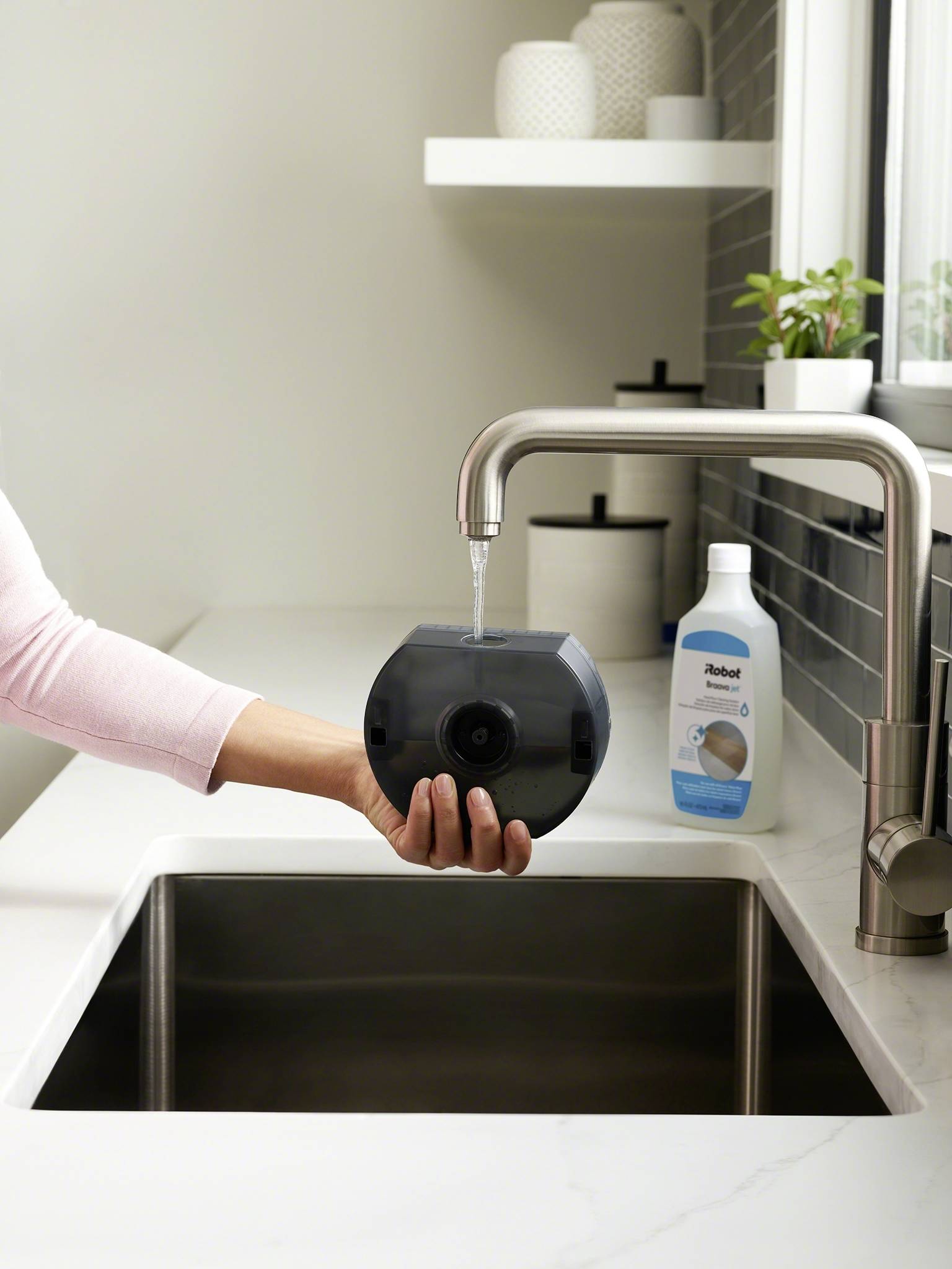 Eine Hand hält einen runden, dunklen Gegenstand unter einen fließenden Wasserhahn über einem Waschbecken, daneben steht Reinigungsmittel.