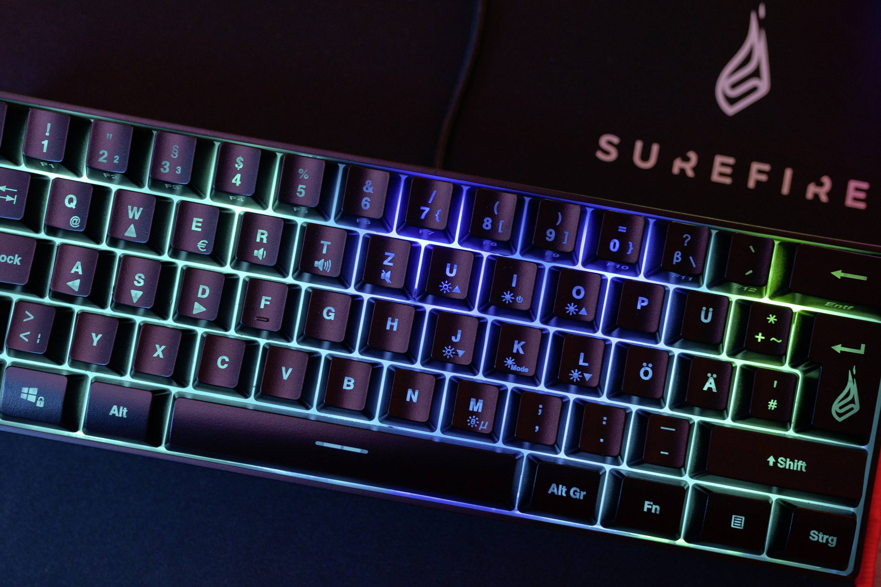 Leuchtende Gaming-Tastatur mit mehrfarbigem LED-Licht und deutschem Layout, Hintergrund zeigt 'Surefire'-Logo.