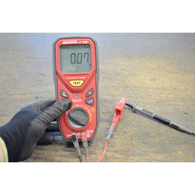 Ein rotes Multimeter zeigt '0.07' an. Eine behandschuhte Hand hält das Gerät, das an zwei Kabel mit Klemmen angeschlossen ist.