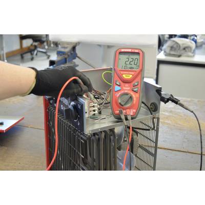 Eine Hand mit schwarzem Handschuh hält ein Multimeter, das an ein elektrisches Gerät angeschlossen ist. Das Display zeigt '220' Volt.