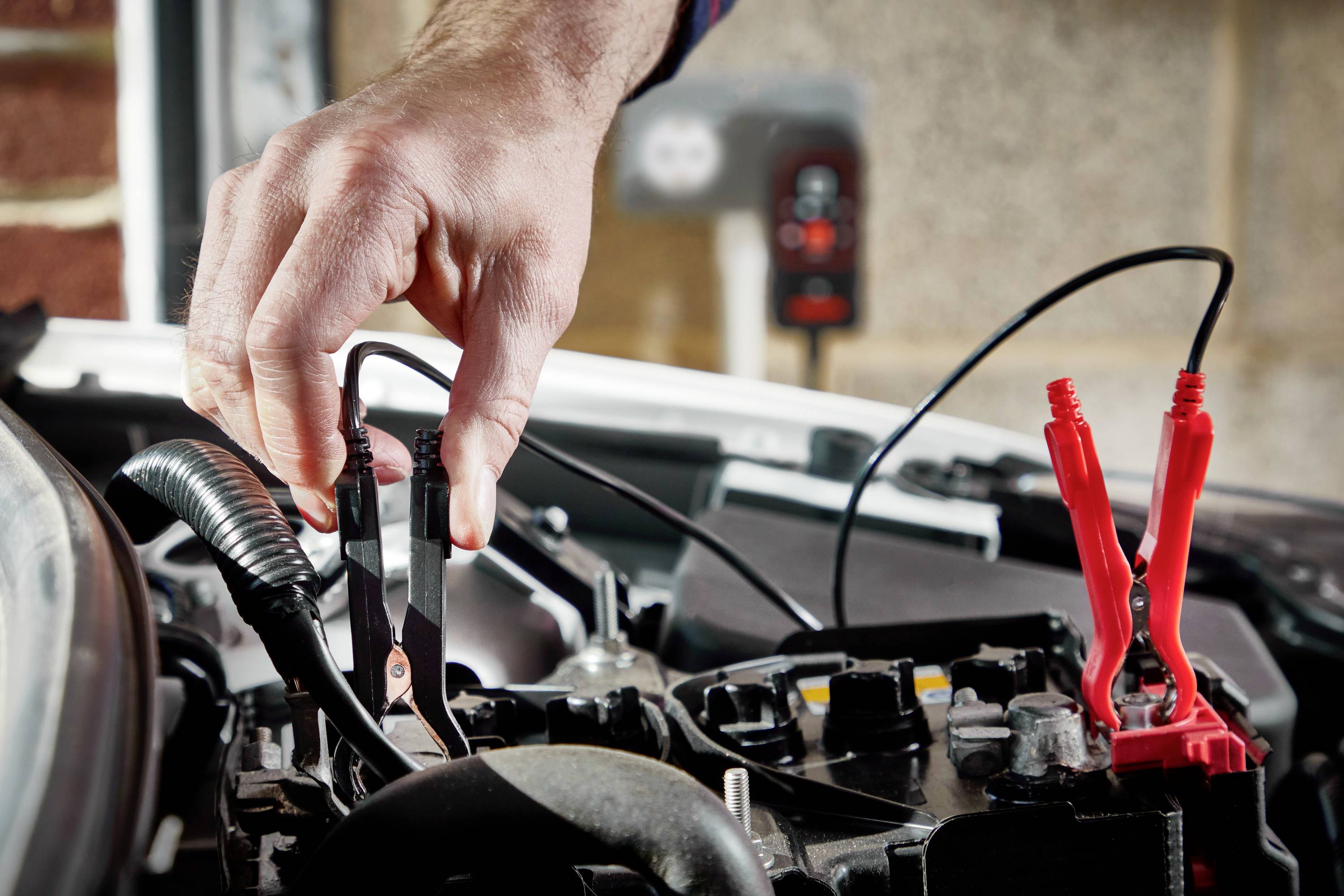 Eine Hand schließt ein schwarzes Überbrückungskabel an die Batterie eines Autos an. Rechts im Bild ist ein rotes Kabel angeschlossen.