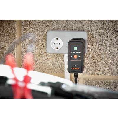 Ein Batterieladegerät von Osram ist an eine Wandsteckdose angeschlossen. Im Vordergrund sind unscharfe rote Klemmen zu sehen.