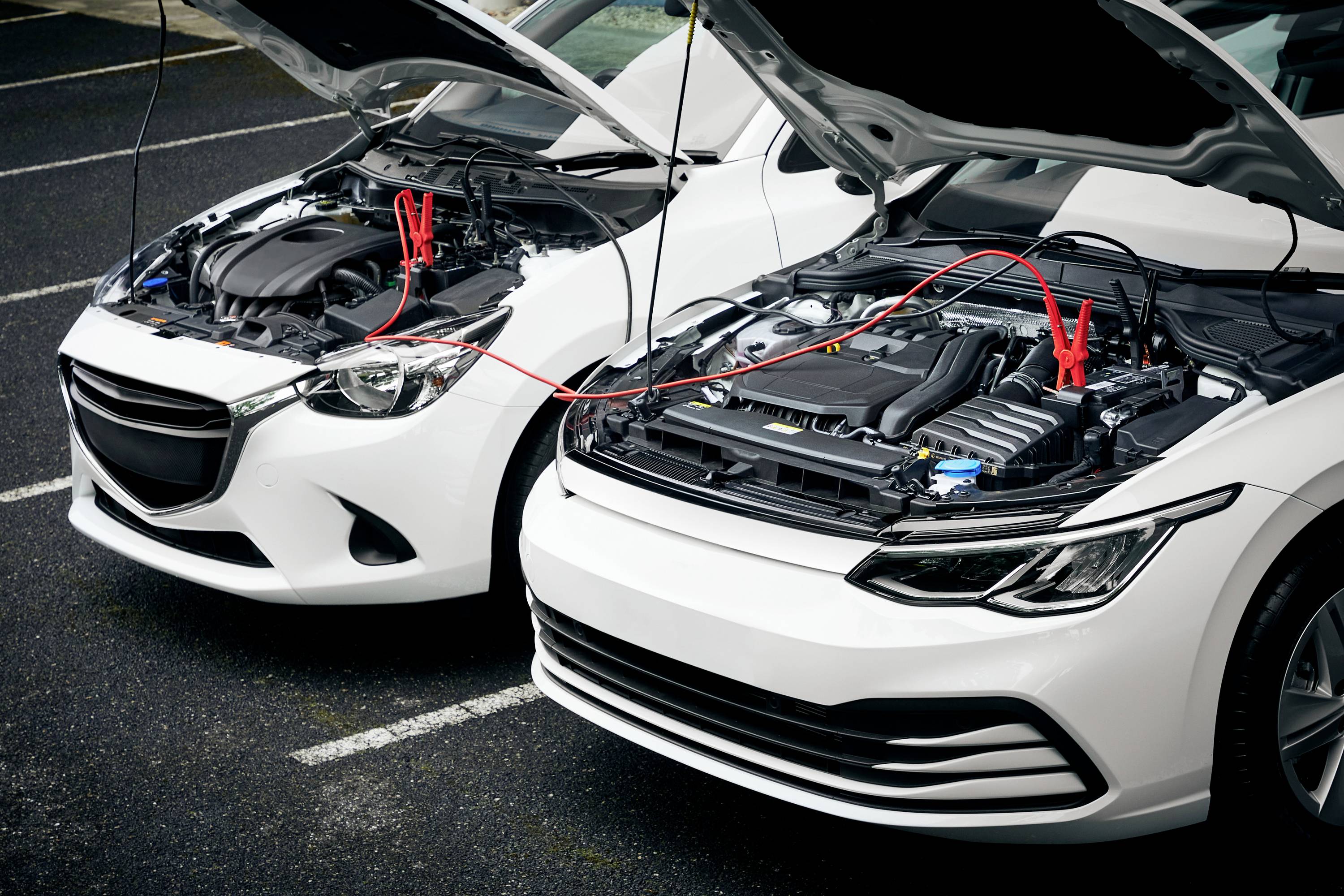 Zwei weiße Autos stehen nebeneinander mit geöffneten Motorhauben. Überbrückungskabel verbinden die Batterien für Starthilfe.