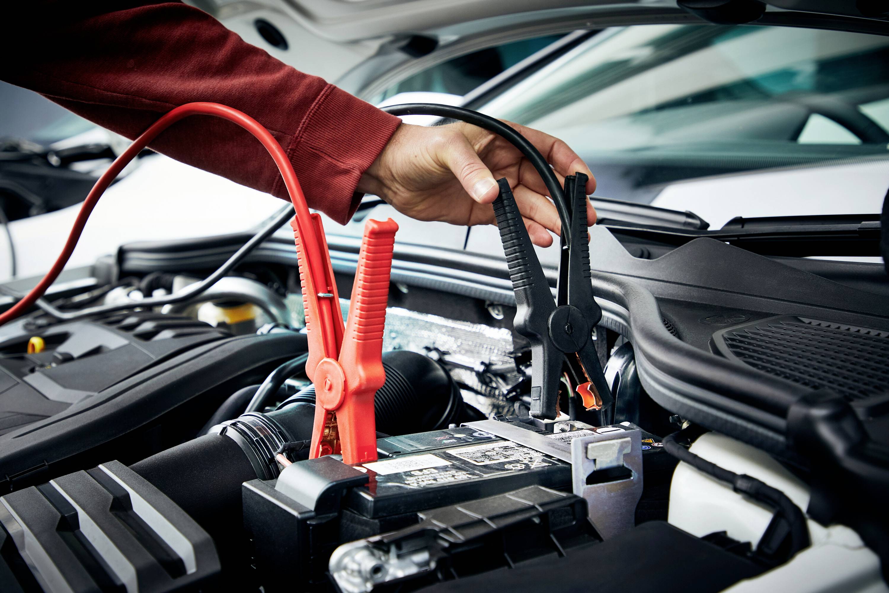 Eine Hand verbindet Starthilfekabel an einer Autobatterie im Motorraum, um ein Fahrzeug zu starten.