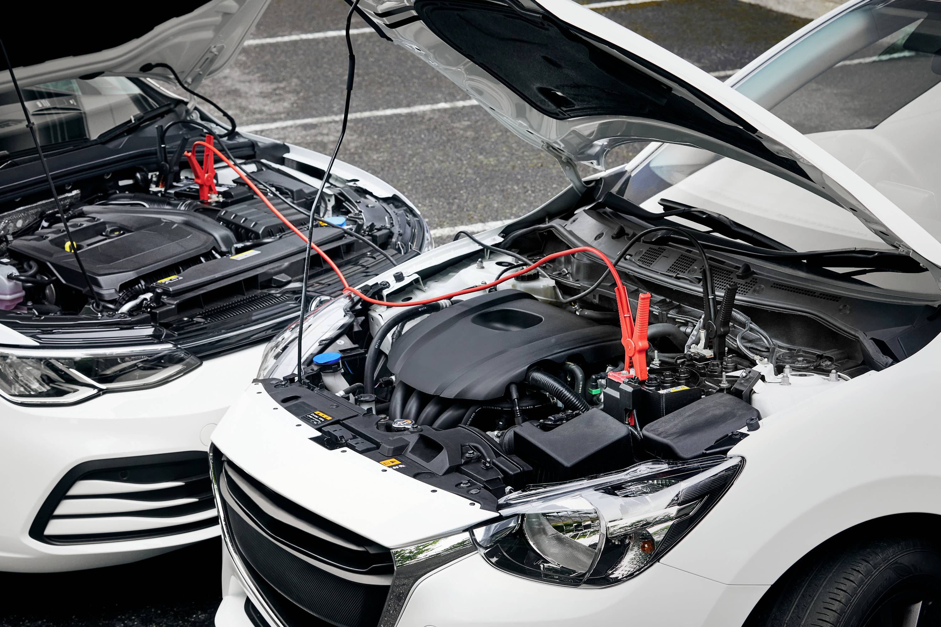 Zwei Autos mit geöffneten Motorhauben sind zu sehen. Überbrückungskabel verbinden die Batterien, offensichtlich für Starthilfe.