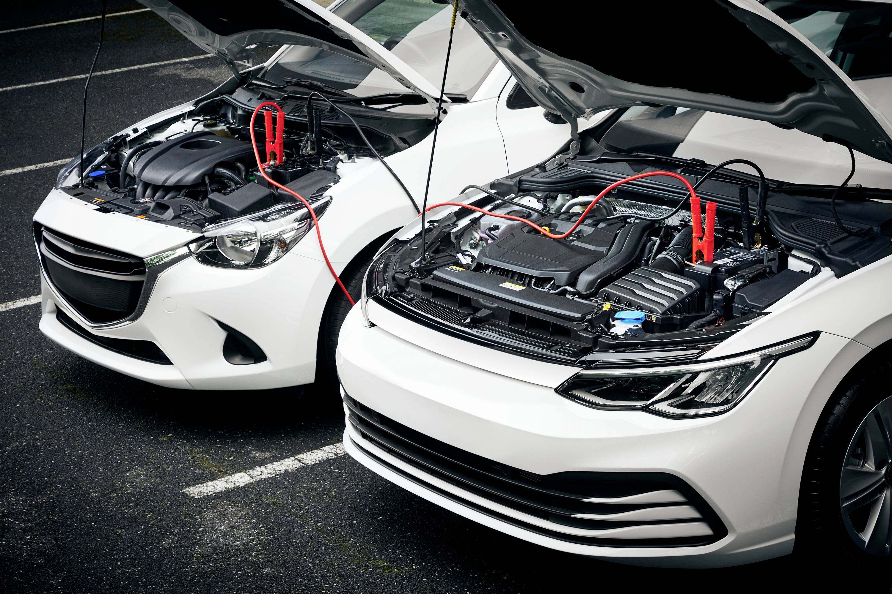 'Zwei Autos mit geöffneter Motorhaube auf einem Parkplatz. Überbrückungskabel verbinden die Batterien, um Starthilfe zu geben.'