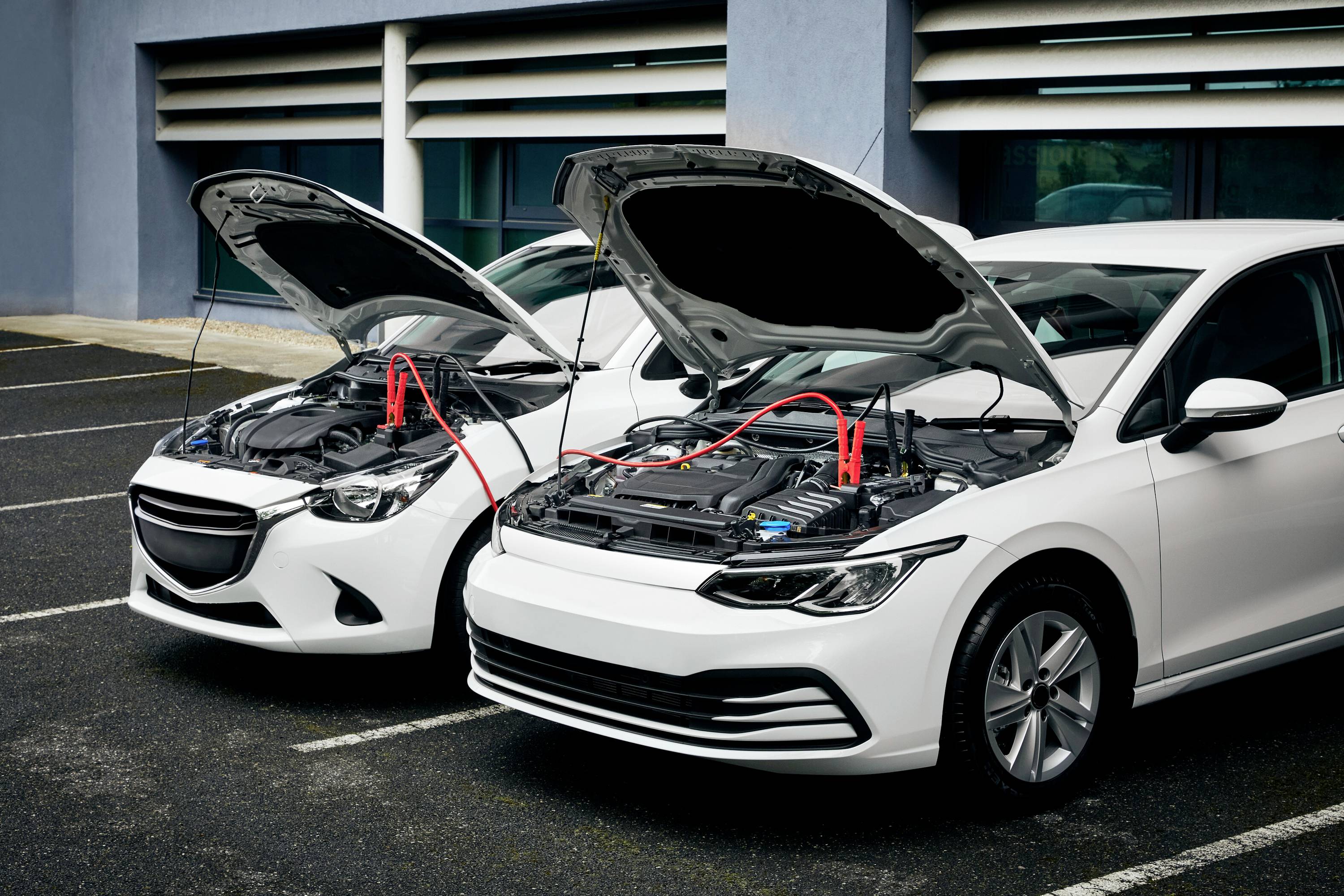 Zwei geparkte Autos mit offenen Motorhauben, verbunden durch ein Starthilfekabel, das den Batteriewechsel unterstützt.