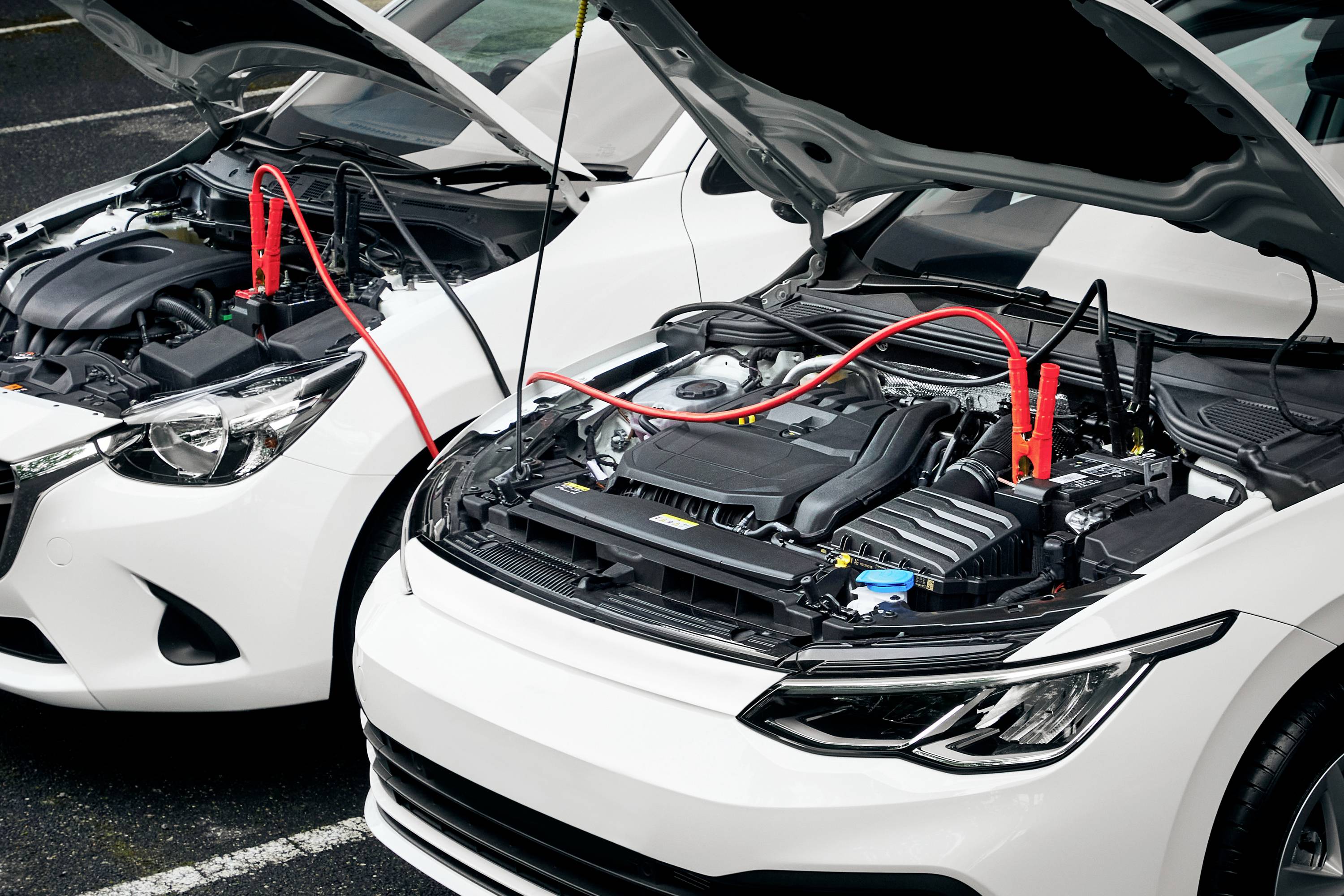 Zwei offene Motorhauben nebeneinander geparkter Autos. Rote Starthilfekabel verbinden die Batterien, um Starthilfe zu geben.