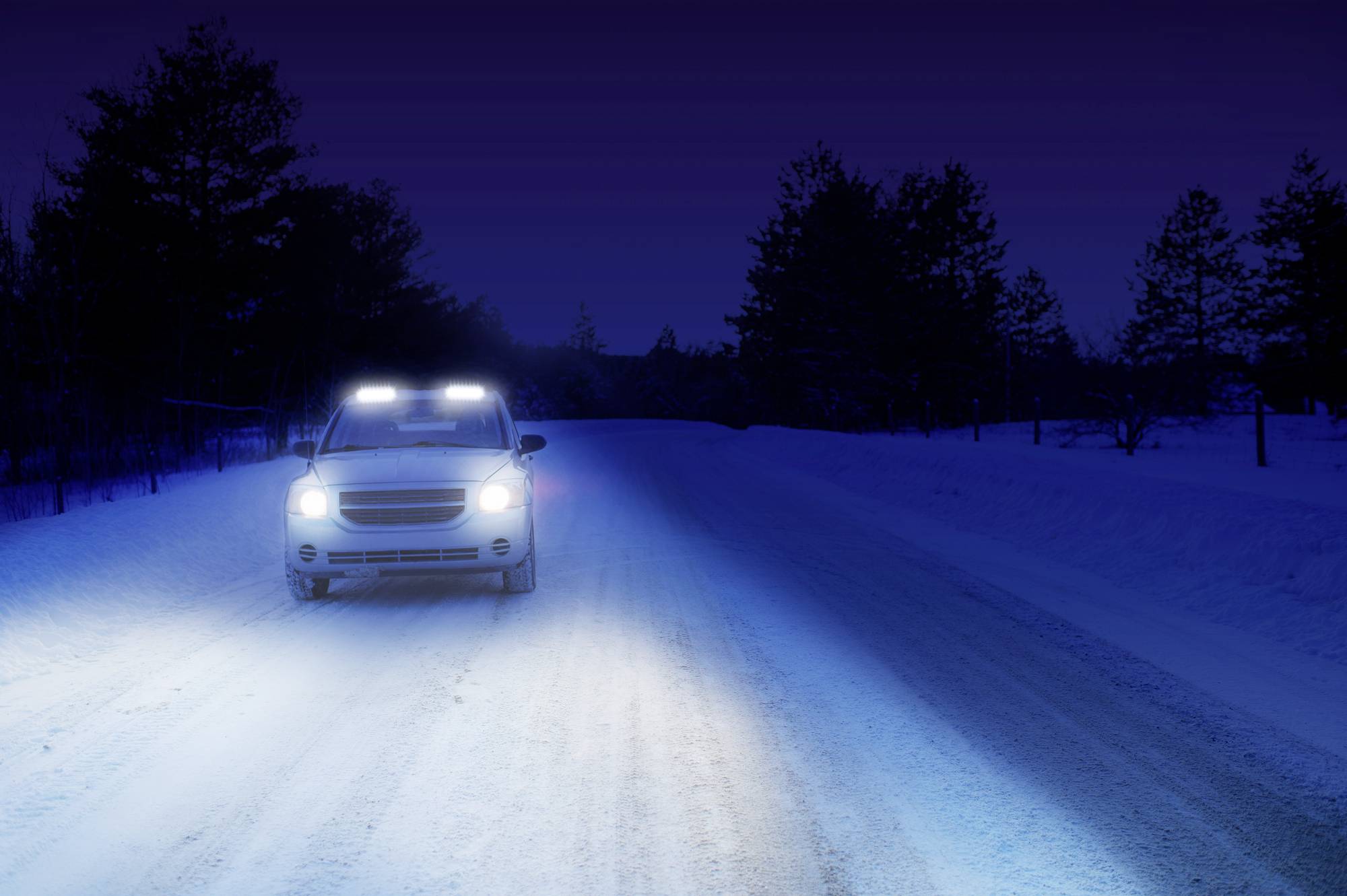 Ein Auto fährt auf einer schneebedeckten Straße bei Nacht. Die Scheinwerfer leuchten stark, Bäume sind im Hintergrund zu sehen.