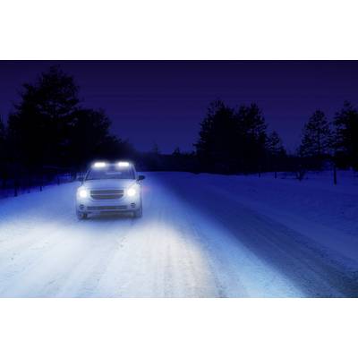 Ein Auto fährt auf einer schneebedeckten Straße bei Nacht. Die Scheinwerfer leuchten stark, Bäume sind im Hintergrund zu sehen.