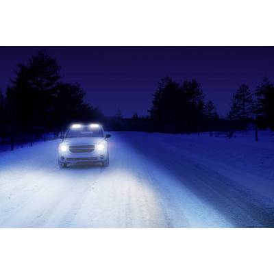 Ein weißes Auto fährt nachts auf einer verschneiten Straße mit eingeschalteten Scheinwerfern, umgeben von schneebedeckten Bäumen.