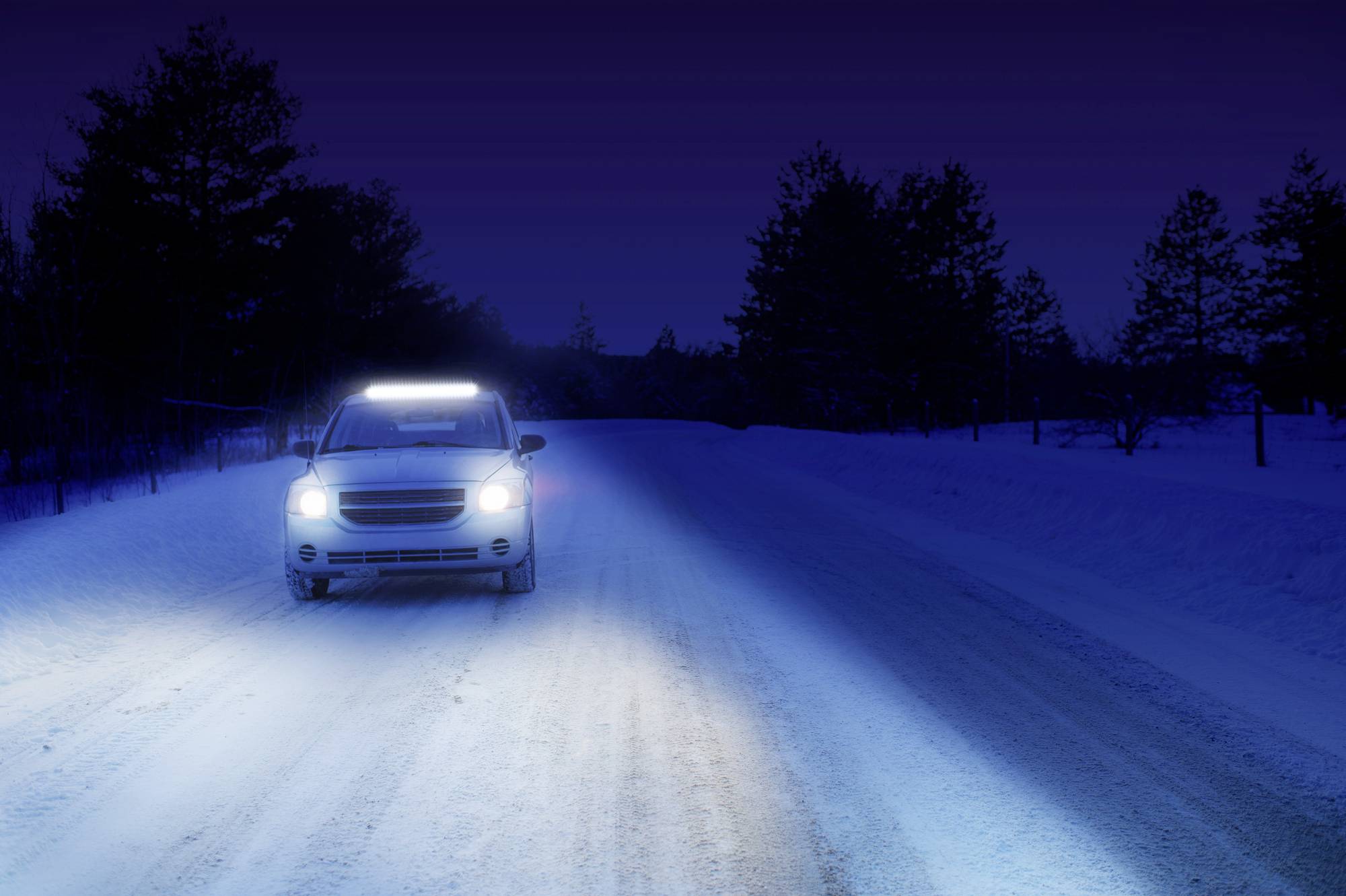 Ein Auto mit eingeschaltetem Fernlicht fährt nachts auf einer schneebedeckten Straße. Bäume säumen die dunkle Umgebung.