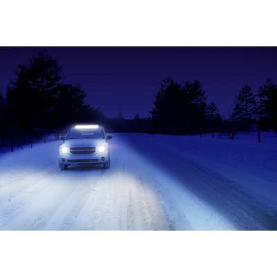 Ein Auto mit eingeschaltetem Fernlicht fährt nachts auf einer schneebedeckten Straße. Bäume säumen die dunkle Umgebung.