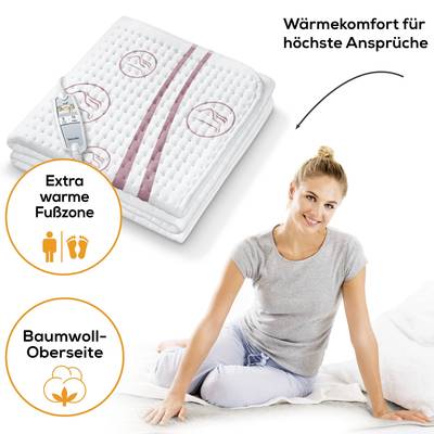 Eine Frau sitzt lächelnd auf einem Bett mit einem Heizkissen. Links sind Symbole für 'Extra warme Fußzone' und 'Baumwoll-Oberseite'. Text: 'Wärmekomfort für höchste Ansprüche'.