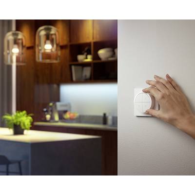 Eine Hand dreht einen smarten Lichtdimmer an der Wand in einem gemütlichen Wohnzimmer mit Küchenbereich im Hintergrund.