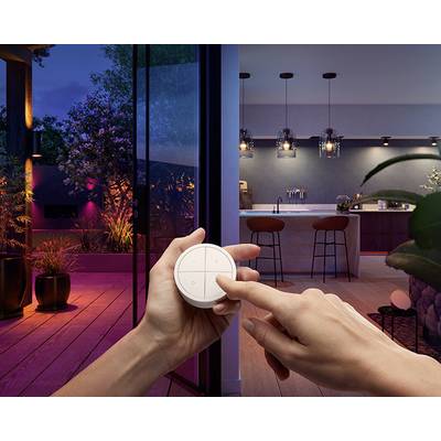 Eine Hand drückt auf eine Fernbedienung in einem modernen Wohnraum; im Hintergrund ein beleuchteter Außenbereich und eine Küche.