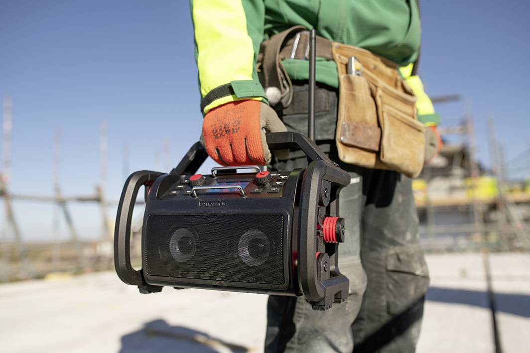 Person in Arbeitskleidung hält ein tragbares Radio auf einer Baustelle. Im Hintergrund sind Baugerüste und ein klarer Himmel zu sehen.