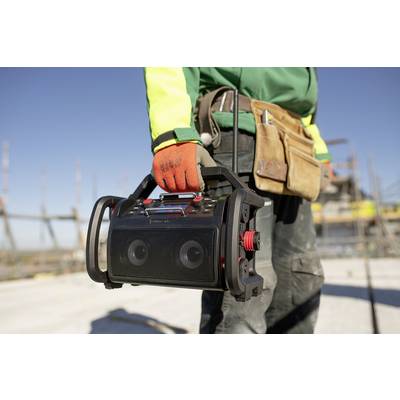 Person in Arbeitskleidung hält ein tragbares Radio auf einer Baustelle. Im Hintergrund sind Baugerüste und ein klarer Himmel zu sehen.
