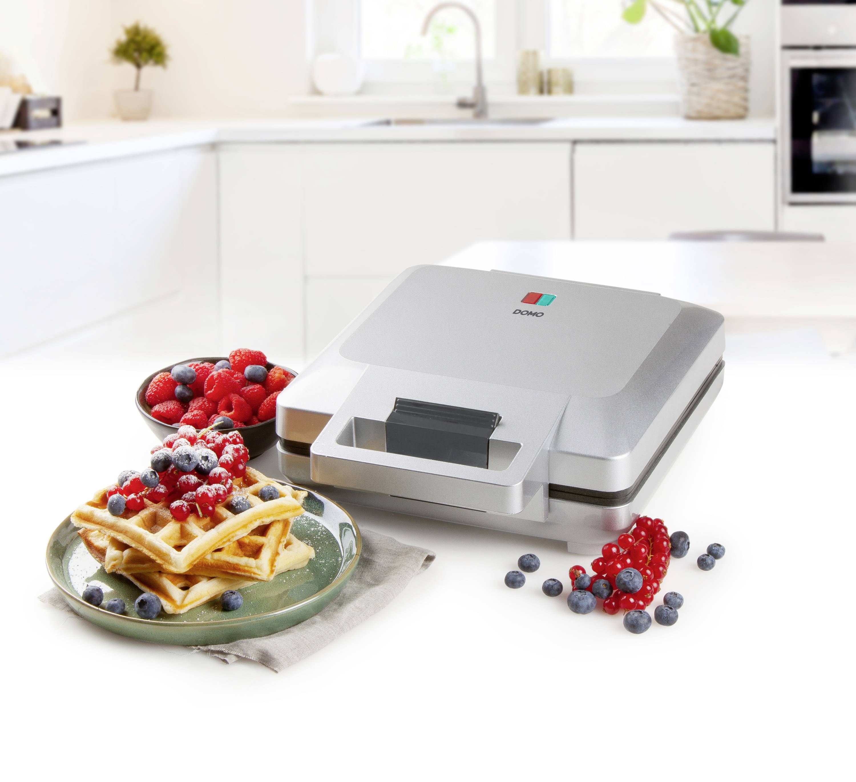 Ein Waffeleisen auf einer Küchenarbeitsplatte. Daneben befinden sich frische Waffeln, garniert mit Beeren. Hintergrund zeigt eine helle, moderne Küche.