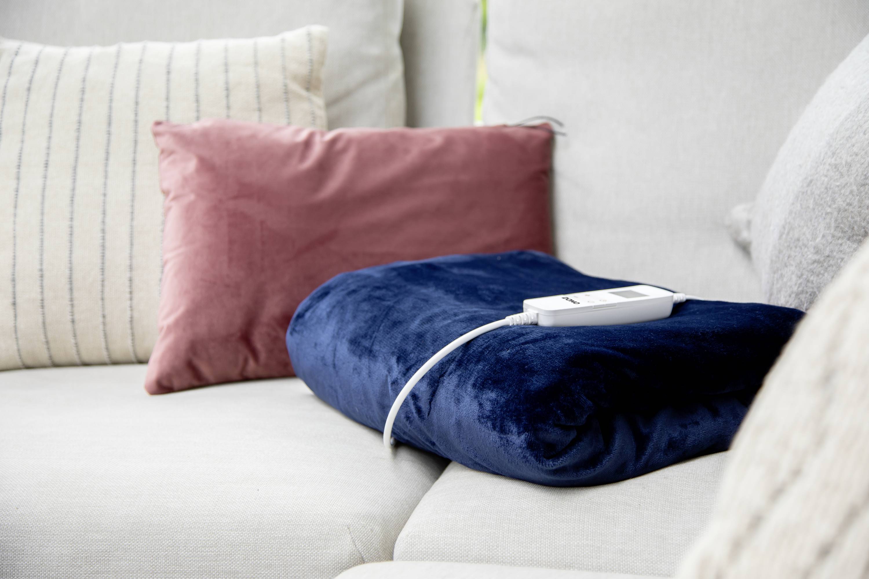 Dunkelblaues Heizkissen mit Kabel auf hellgrauem Sofa, umgeben von farbigen Kissen.