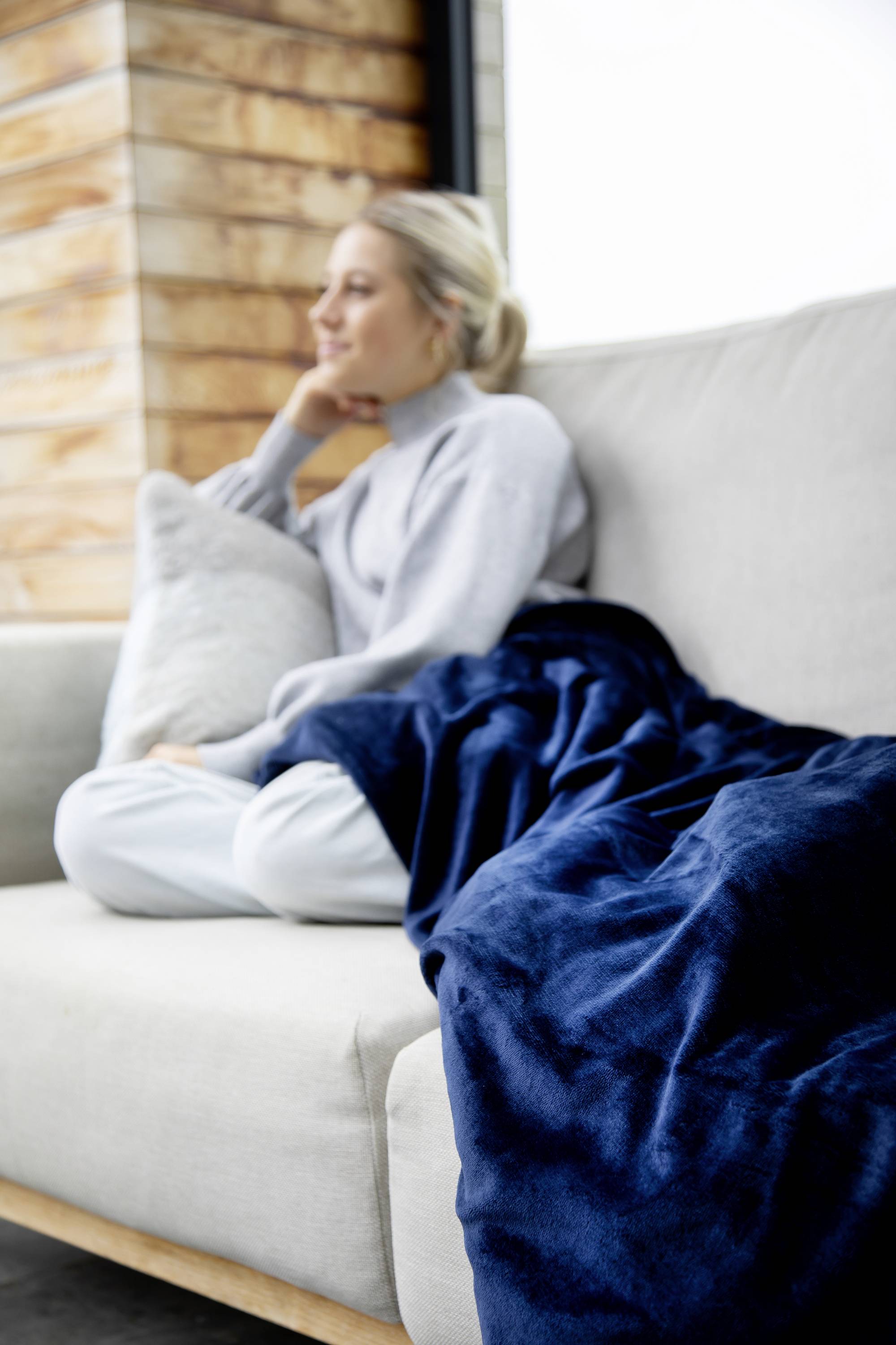 Eine Frau sitzt entspannt auf einem Sofa, umgeben von einem weichen blauen Kissen, und schaut gedankenverloren ins Leere.