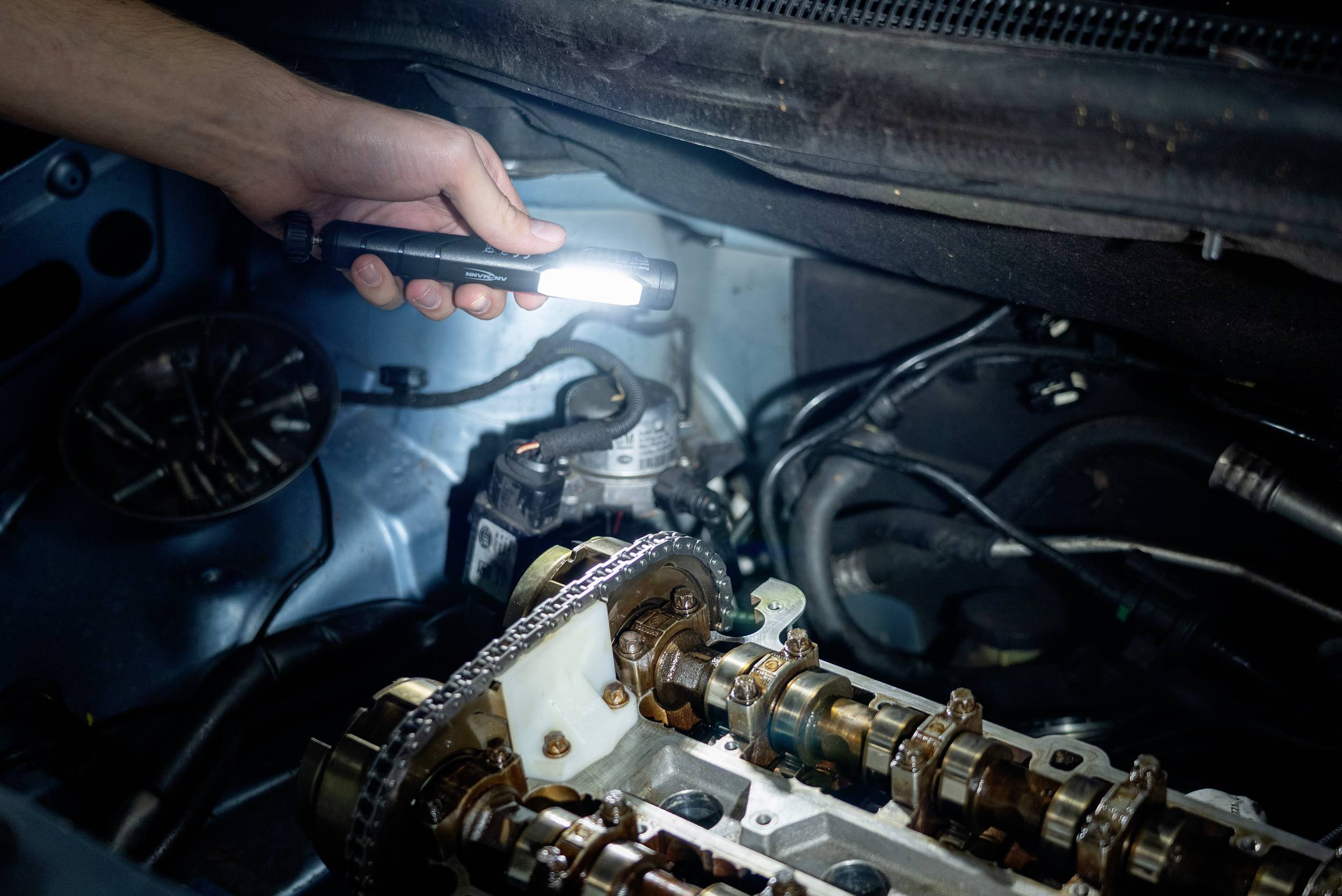 Person überprüft Autobeleuchtung im Motorraum, hält eine Taschenlampe. Sicht auf Motorkomponenten. Wartungsarbeiten werden durchgeführt.